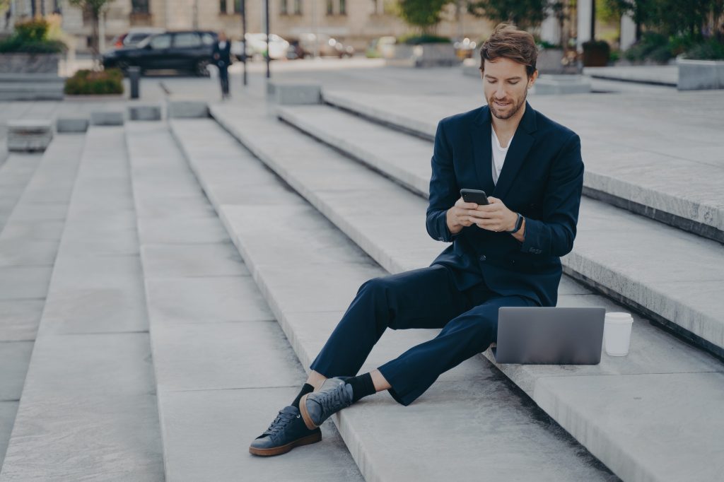 Young confident man in stylish suit using smartphone while sitting on steps outside with laptop